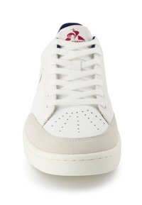 Witte sneaker met geperforeerde neus, lichtbeige suède accenten, witte veters en een logo patch op de tong in rood en marineblauw.