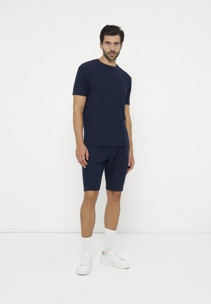 Mand, der står iført en marineblå kortærmet skjorte og shorts, hvide sokker og hvide sneakers mod en ensfarvet hvid baggrund.