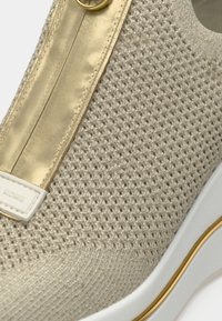 Beigen värinen neulotun mallinen tennari, jossa on kultainen satiinikoriste, teksturoitu pinta ja valkoinen kumipohja kultaisten yksityiskohtien kera. Logolla varustettu sivu.