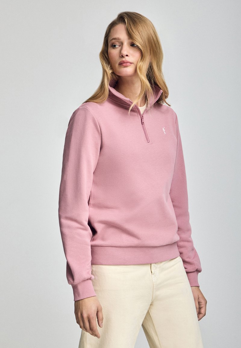 Ljust rosa sweatshirt med halvvinddrag och ribbade ärmar och nederkant. Diskret logotyp på bröstet. Bärs med krämfärgade byxor.