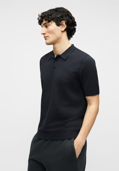 Νέος άντρας με κοντά σγουρά μαλλιά, φορώντας μαύρο πόλο με κοντά μανίκια και μαύρο παντελόνι, με τα χέρια στις τσέπες, κοιτάζει προς τα πλάγια.