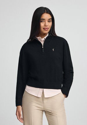 Pull-over noir avec un zip quart, bord côtes en bas et aux poignets. Il présente un petit logo sur la poitrine et est porté par-dessus une chemise à carreaux légère.