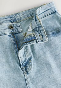 Svijetloplave traperice su slim fit dizajna. Imaju prednji patentni zatvarač, mesingasti gumb, te izbljeđene dijelove s žutim šavovima.
