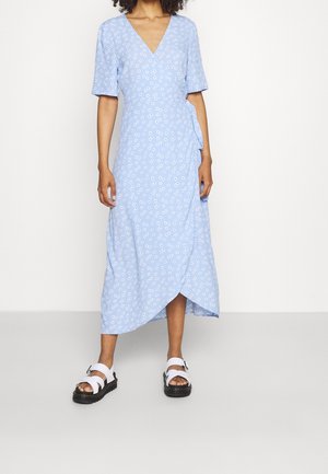 Kobieta ubrana w jasnoniebieską kwiecistą sukienkę kopertową z krótkimi rękawami i białe sandały z czarnymi podeszwami, stojąca na tle białego tła.