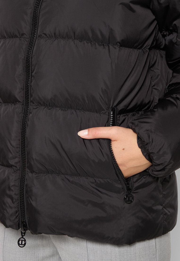 Schwarze Daunenjacke mit Reißverschluss, die über gesteppten Abschnitte, eine seitliche Tasche und elastische Bündchen für einen engen Sitz verfügt.