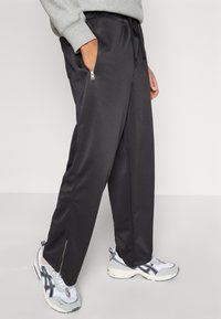 Schwarze Sporthosen mit einem Kordelzugbund, seitlichen Reißverschlusstaschen und einer glatten Textur, kombiniert mit grau-weißen Sneakers.