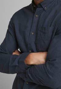 Tmav�ě modrá košile na knoflíky s texturovanou tkaninou, dlouhými rukávy a náprsní kapsou. Obsahuje malé zlaté knoflíky podél zapínání.