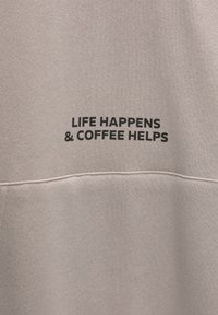 Tissu beige avec un texte noir indiquant « LIFE HAPPENS & COFFEE HELPS » au-dessus d'une couture horizontale.