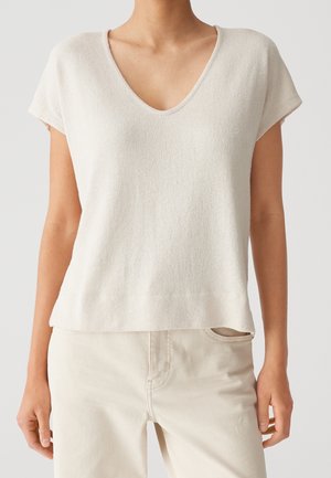 Vrouw draagt een beige gebreide top met korte mouwen en V-hals en lichte broek tegen een effen achtergrond.