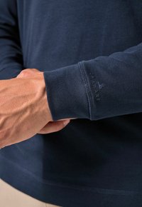 Marineblaues Langarmshirt mit geripptem Bündchen. Der Stoff wirkt glatt und hat ein dezentes Logo am Bündchen. Hände sind im Vordergrund zusammengelegt.