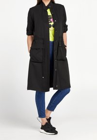 Giacca lunga nera con collo a risvolto, bottoni frontali, due grandi tasche frontali e coulisse. Indossata sopra una maglietta grafica verde brillante e jeans.