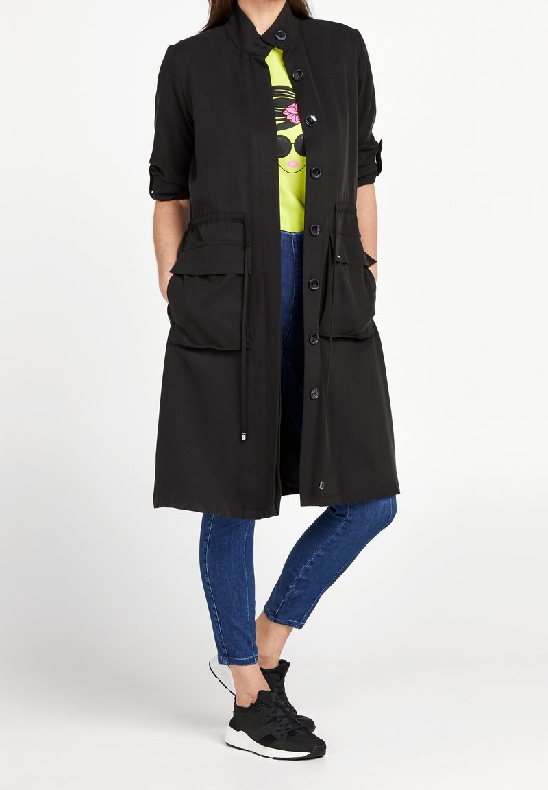 Giacca lunga nera con collo a risvolto, bottoni frontali, due grandi tasche frontali e coulisse. Indossata sopra una maglietta grafica verde brillante e jeans.