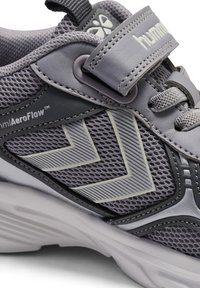 Grå sportssko med meshoverdel, der har teksturerede detaljer, en velcrolukning og en hvid sål. Designet inkluderer vinkelformede mønstre for ekstra detalje.