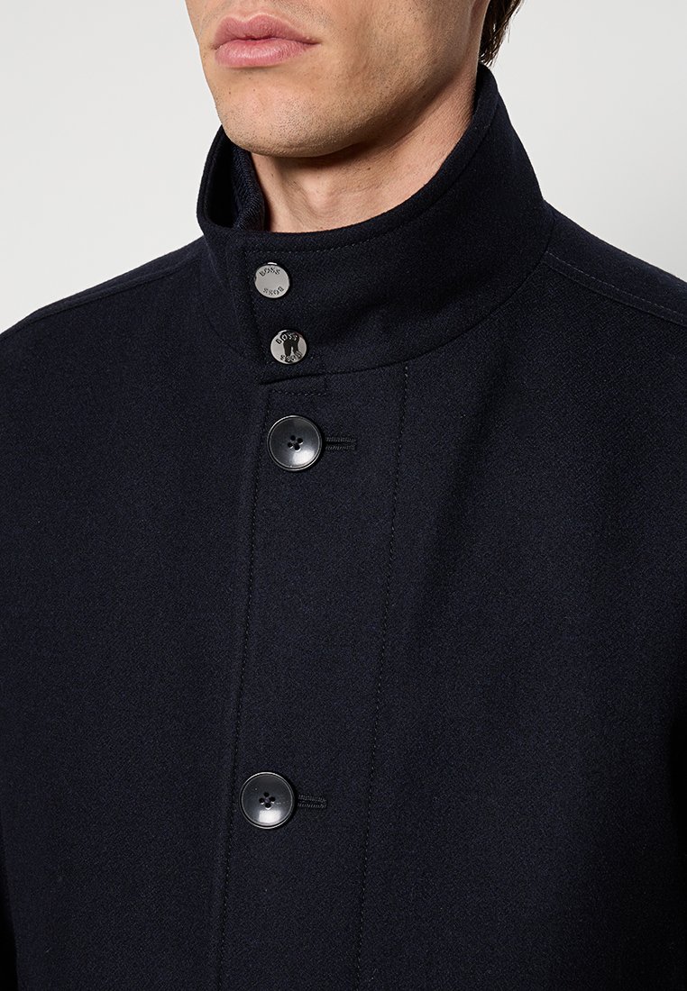 Cappotto di lana blu navy con colletto alto, dotato di bottoni a pressione argentati e bottoni neri. Tessuto testurizzato con un design pulito e su misura.