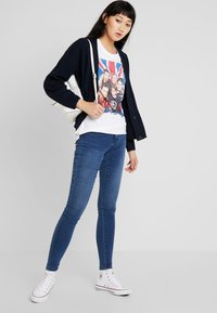 Model trägt eine marineblaue Strickjacke über einem weißen T-Shirt mit einem Union Jack Design, kombiniert mit blauen Skinny Jeans und weißen Sneakers.