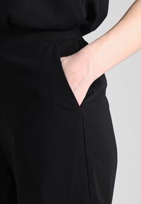 Pantalon noir sur mesure avec une texture lisse, doté de poches latérales et d'une taille ajustée, présentant un design épuré et minimaliste.