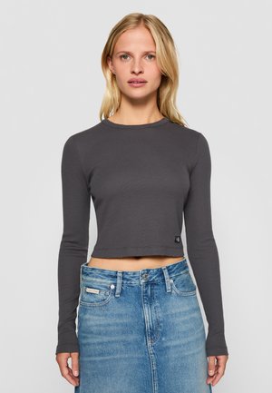 Halliltud hall ribakujuline pika varrukaga cropped topp, millel on ümmargune kaelus ja topitud serv. Kombineeritud kõrge talje ja helesinise denim seeliku ning korsetiga.