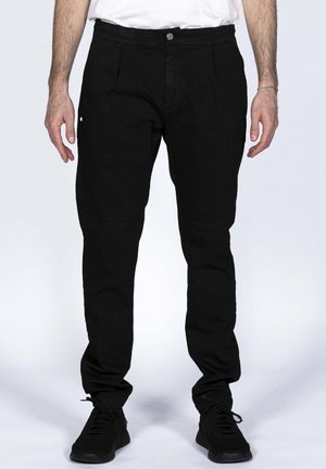 Mann trägt schwarze Slim-Fit-Hosen und schwarze Sneakers, steht mit entspannten Armen seitlich vor einem schlichten Hintergrund.