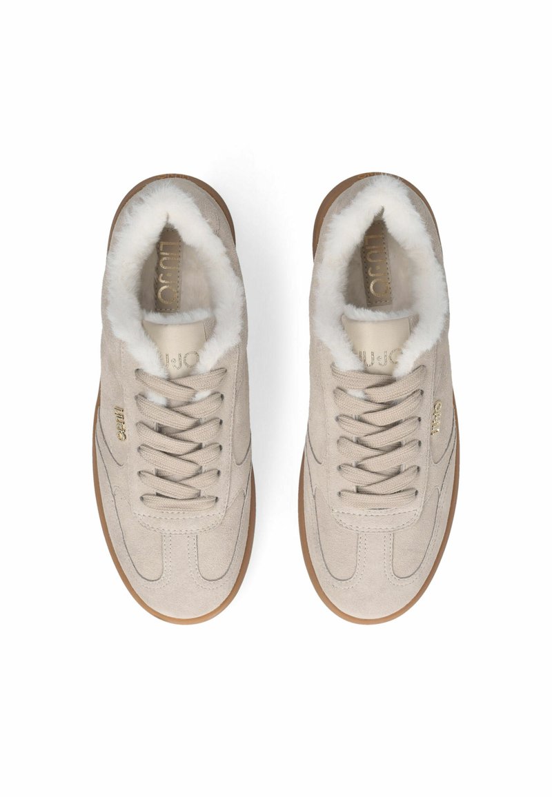 Beige Wildlederschuhe mit einem weichen weißen Futter aus Fleece, flachen Schnürsenkeln, strukturierten Details und einer tanfarbenen Gummisohle. Goldene Markenkennzeichnung an der Seite.