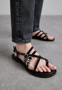 Sandali neri piatti con cinturini incrociati, realizzati in materiale texturizzato, con accentuazioni metalliche e un design semplice e aperto sulla punta.
