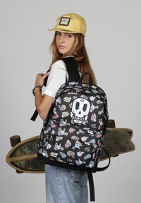 Mochila negra con estampados gráficos coloridos, incluyendo un diseño de calavera. Presenta correas ajustables y un bolsillo frontal. Llevada sobre una camiseta blanca.