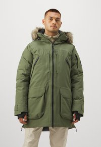 Parka verde con cappuccio foderato in pelliccia, presenta diverse tasche frontali, polsini regolabili e una zip a lunghezza totale. Materiale liscio e resistente.