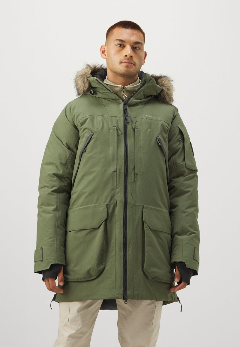 Parka verde con cappuccio foderato in pelliccia, presenta diverse tasche frontali, polsini regolabili e una zip a lunghezza totale. Materiale liscio e resistente.