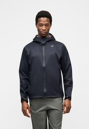 Muž v tmavomodrej nepremokavej bunde s kapucňou a predným zipsom a sivými nohavicami, stojaci na bielom pozadí.