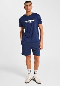 Dunkelblaues Baumwoll-T-Shirt mit Frontlogo, kombiniert mit dunkelblauen Shorts. Weiße und schwarze gestreifte Socken und beigefarbene Sneakers runden das Outfit ab.