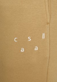 Tecido de algodão em tom cáqui com letras brancas bordadas a dizer "casll." A textura é suave com uma ligeira elasticidade. Não há outros detalhes visíveis.