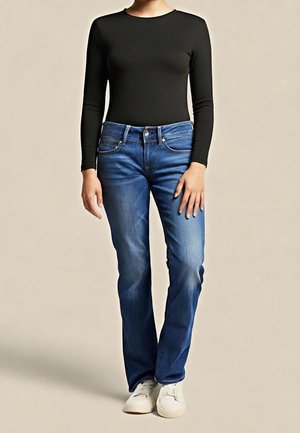 Personne portant un haut noir ajusté à manches longues, un jean bleu droit et des baskets blanches, debout devant un fond uni.