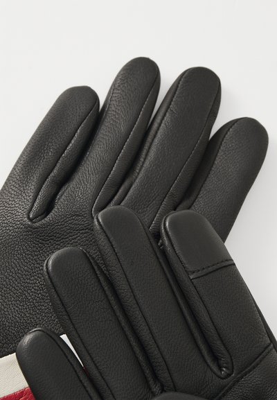 Gants en cuir noir avec une surface texturée, dotés de coutures distinctes et d'accents rembourrés sur les doigts, disposés sur un fond clair.