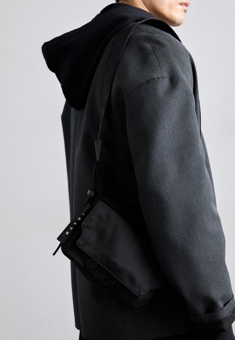Sac à bandoulière en tissu noir avec une texture lisse, doté d'une sangle avec logo, de fermetures magnétiques et d'une forme rectangulaire élégante.