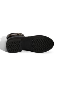 Zwarte leren laars met een platte rubberen zool met een zigzagpatroon. Gouden hardware accenten bij de enkel. Zachte, gestructureerde bovenkant.