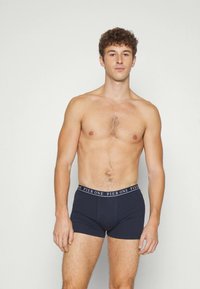 Felsőtest nélkül álló, göndör hajú férfi fekete boxer alsónadrágban, ami a "PIER ONE" logójával díszített derékrészen, sima világos háttér előtt.