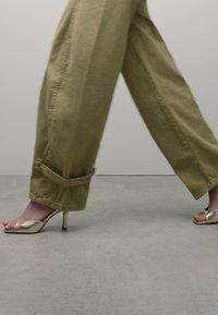 Beige wijde broek met opgerolde manchetten en een glanzende gouden open-teen hoge hak, die een slank design en een soepele textuur benadrukt.