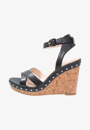 Sandalia de cuña de cuero negro con correa ajustable en el tobillo, base con textura de corcho y tachuelas metálicas decorativas a lo largo de la suela.