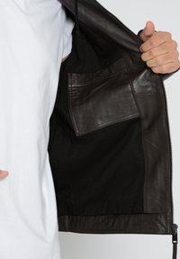 Brun læderjakke med en sort foring, der har en indvendig lomme med en knap og en lynlåslukning nederst.