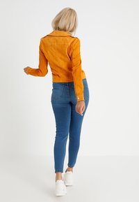 Oranje suède jas gecombineerd met blauwe skinny jeans en witte sneakers. De jas heeft een kraag en een aansluitend ontwerp.