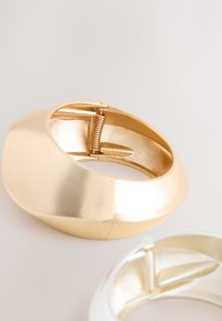 Goldfarbenes Metallarmband mit glatter Oberfläche, ausgestattet mit einem Feder-Scharnierverschluss. Das Design ist oval geformt und hebt ein glänzendes Finish hervor.