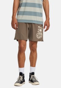 Pantaloni da bagno marroni con una grafica astratta bianca sulla gamba sinistra. Abbinati a sneaker nere alte e calzini bianchi. Camicia a righe sopra.