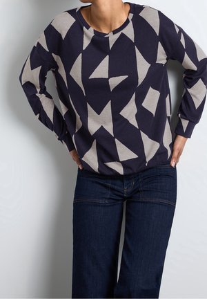 Persoon draagt een marineblauwe trui met beige abstracte geometrische vormen en donkerblauwe spijkerbroek, staand met handen in de zij tegen een effen achtergrond.