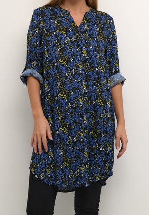 Femme portant une tunique à manches longues avec col, imprimé floral bleu, jaune et blanc, manches retroussées aux coudes, associée à un pantalon noir.