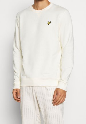 Mężczyzna ubrany w biały sweter crewneck z małym żółtym logo ptaka na piersi oraz białe spodnie z cienkimi pionowymi paskami.