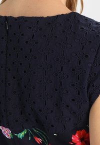 Blusa blu navy con dettagli di pizzo intricati, caratterizzata da ricami floreali nella parte inferiore. Include una chiusura con cerniera discreta sul retro.