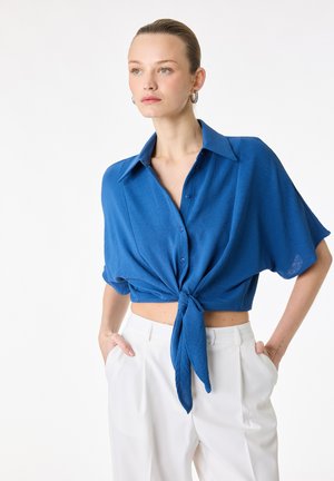 Vrouw die een blauwe, korte mouwen, knoopblouse met strik aan de voorkant draagt en een witte broek met hoge taille, staand met haar handen in de zakken.