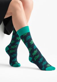 Nohy zkřížené, obuté v tmavě modrých ponožkách se vzorem zelených listů a s jednobarevnými zelenými lemy, špičkami a patami na bílém pozadí.