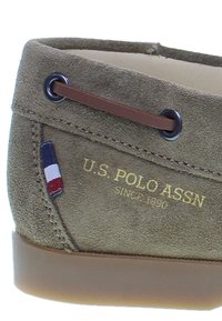 Scarpa in camoscio verde oliva con dettagli in pelle marrone, occhielli rotondi e un piccolo logo tricolore vicino al tallone. Fodera morbida e suola in gomma.