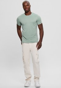 Camiseta de manga corta verde claro con cuello redondo, combinada con pantalones cargo crema que tienen bolsillos laterales. Llevada con zapatillas blancas.