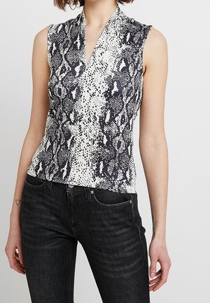 Mouwloze zwart-witte wikkeltop met slangenprint gecombineerd met zwarte hoog getailleerde jeans bij een persoon die staat tegen een effen achtergrond.